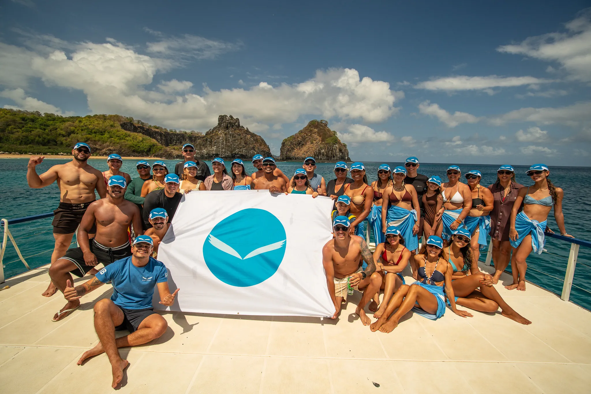 Grupo da RIB Turismo com a bandeira da marca no deck de um barco, diante do Morro Dois Irmãos, em Fernando de Noronha